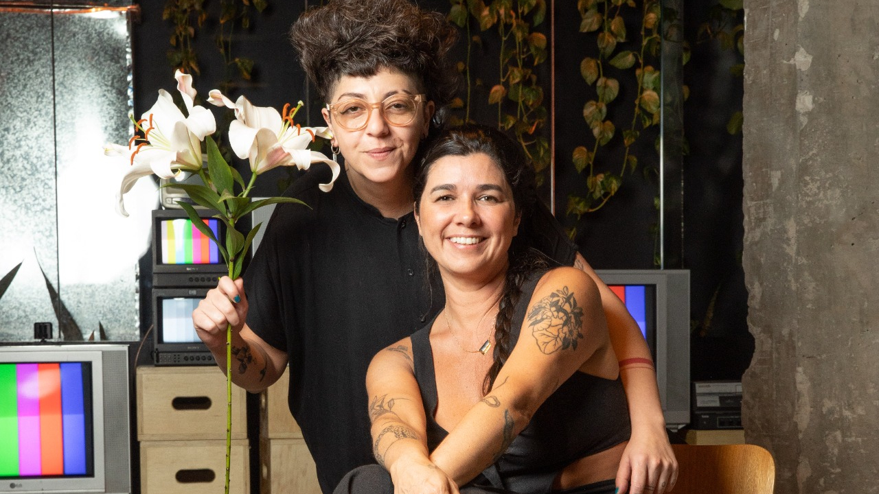 Duas mulheres de preto, uma sentada, outra abaixada segurando um bouquet de copo de leite, abraçada uma a outra.