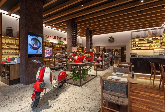 Imagem da loja Casa La Pastina. Do lado direito estão as mesas e cadeiras. No centro tem decorações como uma scooter vermelha. Nas paredes ao fundo estão prateleiras com vinhos.