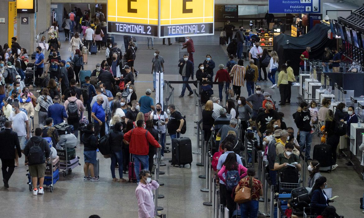 saguão de aeroporto cheio de pessoas com máscaras no rosto segurando suas malas