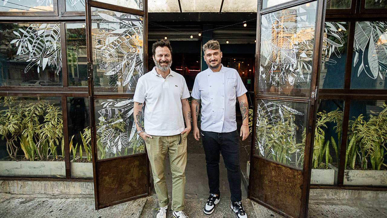 A imagem mostra Alê D'Agostino e Thiago Cerqueira ao lado um do outro. Eles estão na entrada do bar, entre duas portas de vidro abertas.