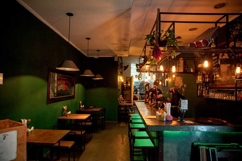 A imagem mostra o interior do bar com mesas e cadeiras à esquerda e um balcão à direita.