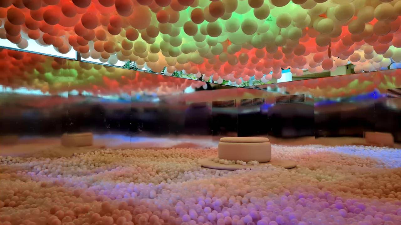 A imagem mostra uma sala ampla com uma piscina de bolinhas coloridas e balões pendurados no teto.