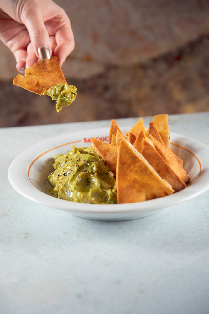 Prato branco com guacamole à esquerda e pedaços de pão no formato retangular à direita. Uma mão segura um pedaço do pão com guacamole.