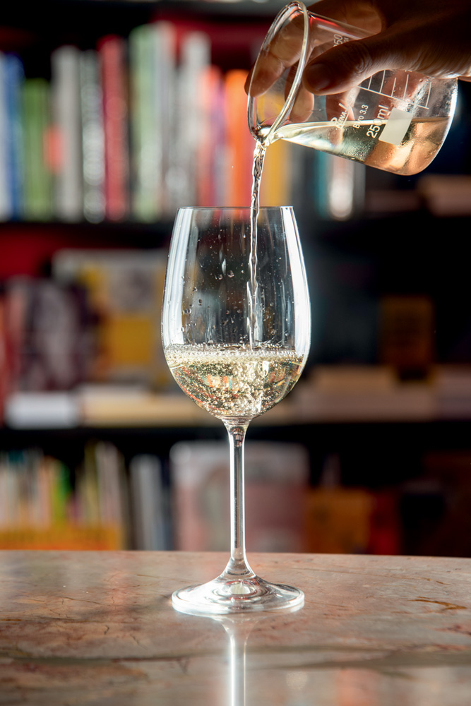 Barouche Vinho branco sendo colocado dentro de uma taça. Ao fundo, prateleiras cheias de livros.