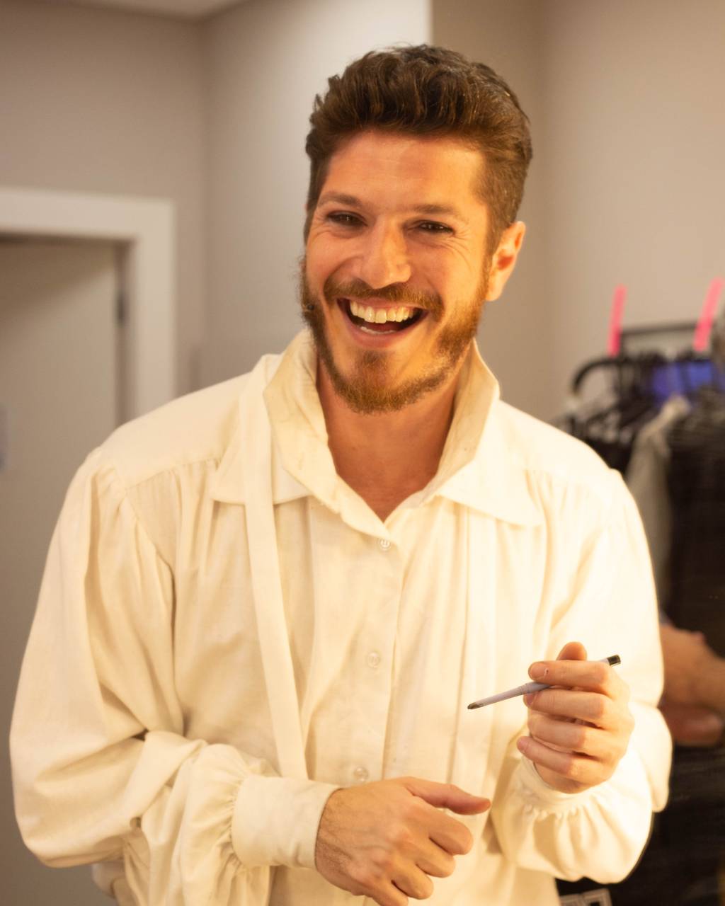 Caio Paduan (Rafa Marques-Divulgação) Homem loiro branco com camisa branca sorri em camarim. Segura uma caneta.