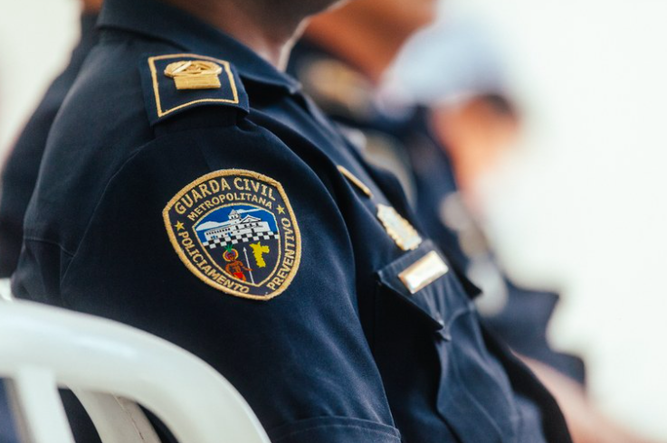 um guarda civil metropolitano sentado em uma cadeira de plástico. a foto foca no distintivo da GCM em seu uniforme.