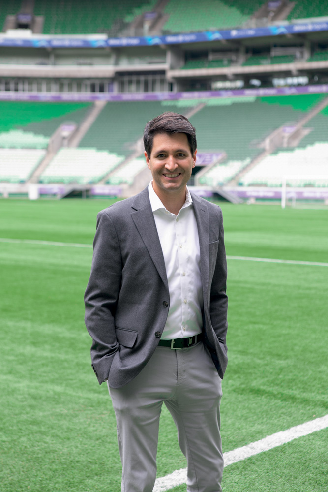 Imagem mostra Cláudio Macedo em campo de futebol, usando blazer, camisa e calça social. Ele sorri para a foto em dia ensolarado