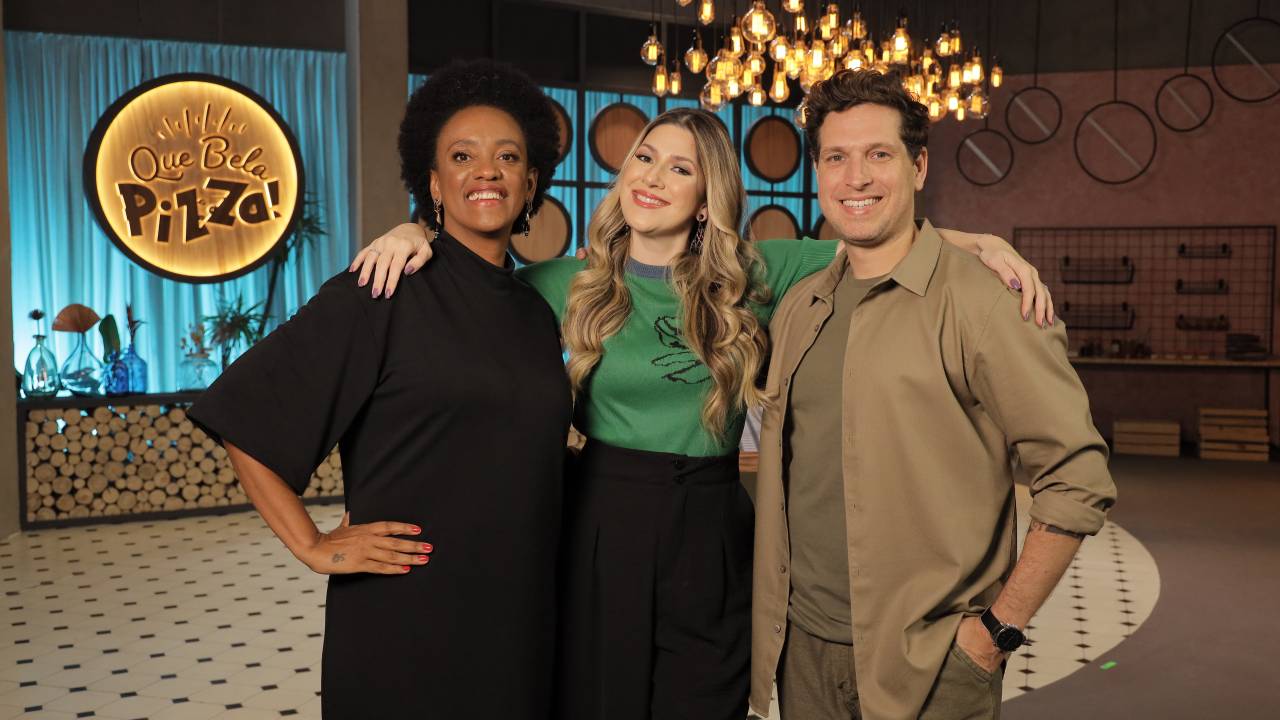 Foto com duas mulheres, à esquerda e ao centro, e um homem abraçados e sorrindo para a foto no estúdio do programa Que bela pizza.