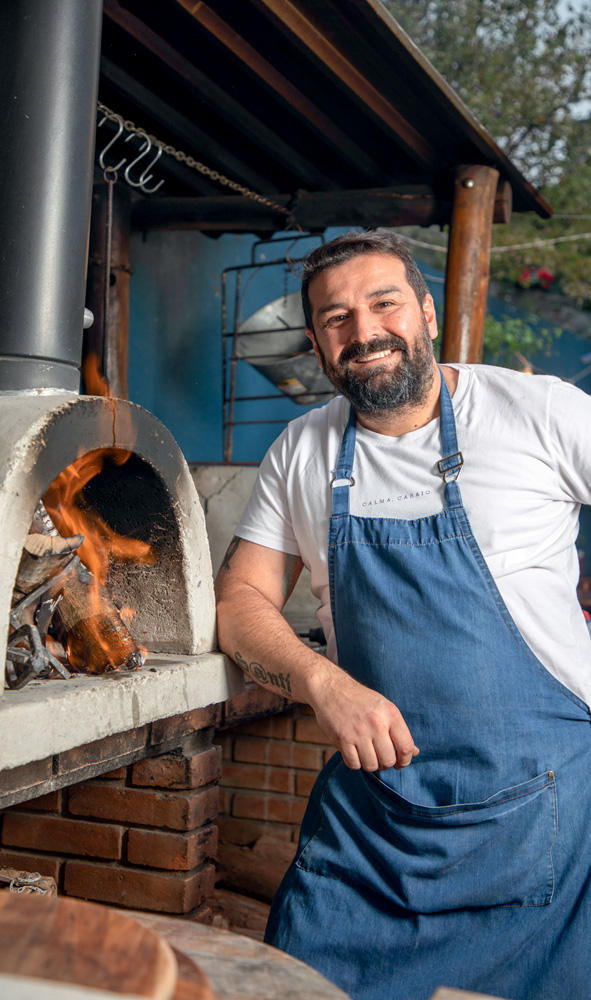 Rotiseria Argentina _ comidinhas A imagem mostra um forno a lenha à esquerda, e ao lado um homem de camiseta branca e avental azul sorrindo.