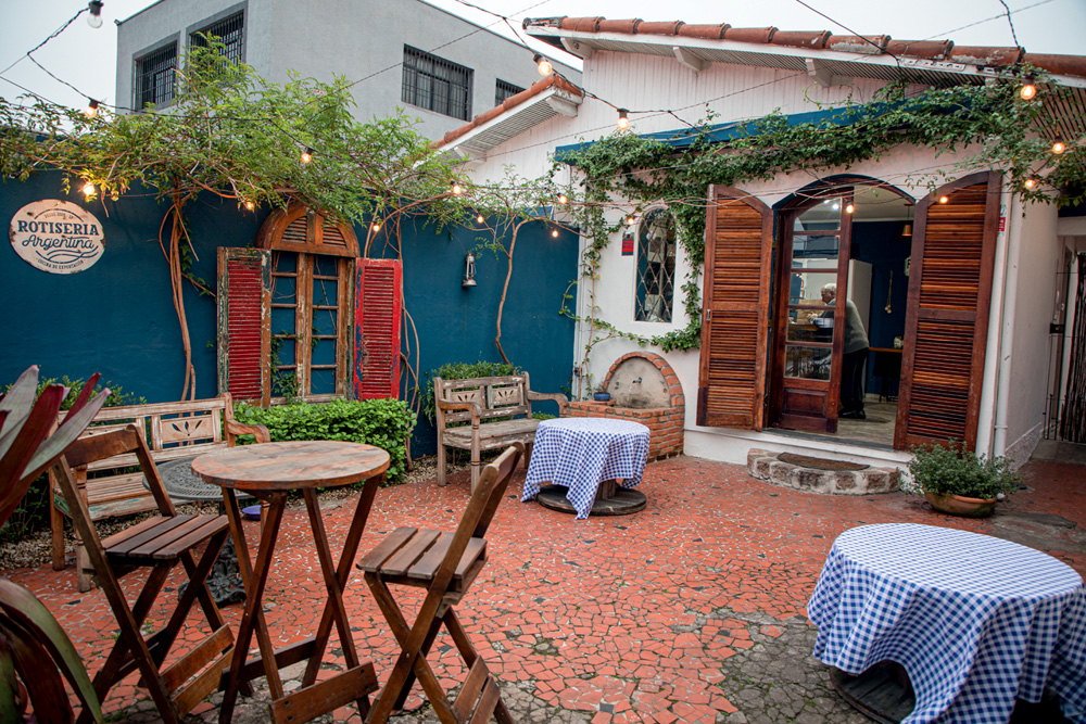 A imagem mostra o quintal com paredes azuis e mesas de madeira da rotisseria.