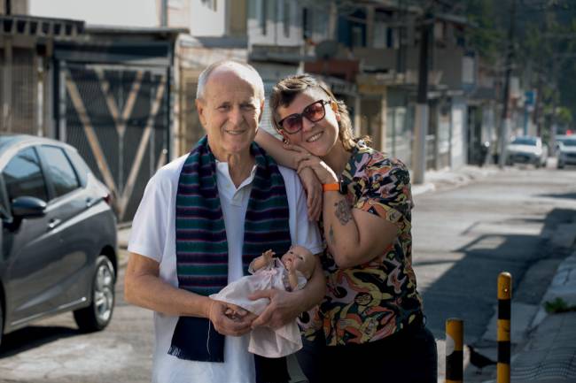 Imagem mostra o pai, Adelchi, segurando uma boneca no colo. Ele usa um cachecol escuro no pescoço. A filha, Marcela, aparece sorrindo, apoiada no ombro do pai. Eles estão em uma rua residencial da Lapa