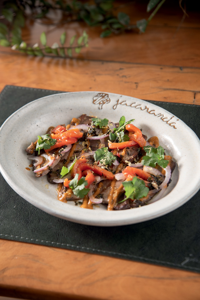 Jacarandá Prato fundo de louça branca sobre jogo americano preto em mesa de madeira com salada de língua com pimentão, cebola e coentro.