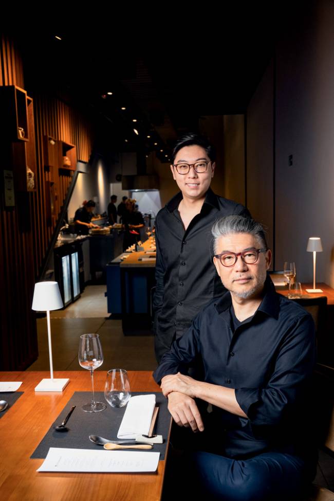jun e tsuyoshi murakami Imagem mostra pai, Tsuyoshi, sentado em mesa de restaurante e filho, Jun, logo atrás. Pai olha sério para a foto e filho sorri