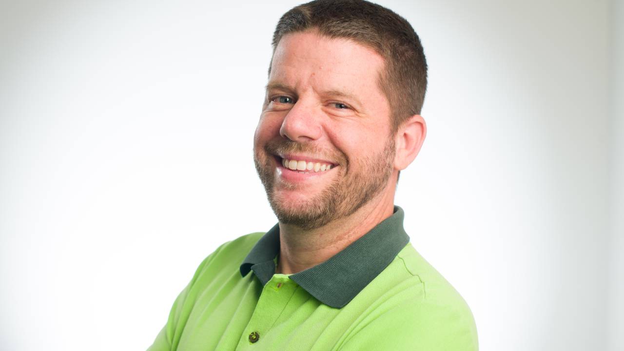 Homem branco com barba e cabelo ralos sorri para a câmera. Posa em fundo branco com camisa polo verde.