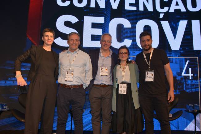 Imagem mostra cinco pessoas reunidas em palco. Na parte de trás, pode-se ler em telão: Convenção Secovi