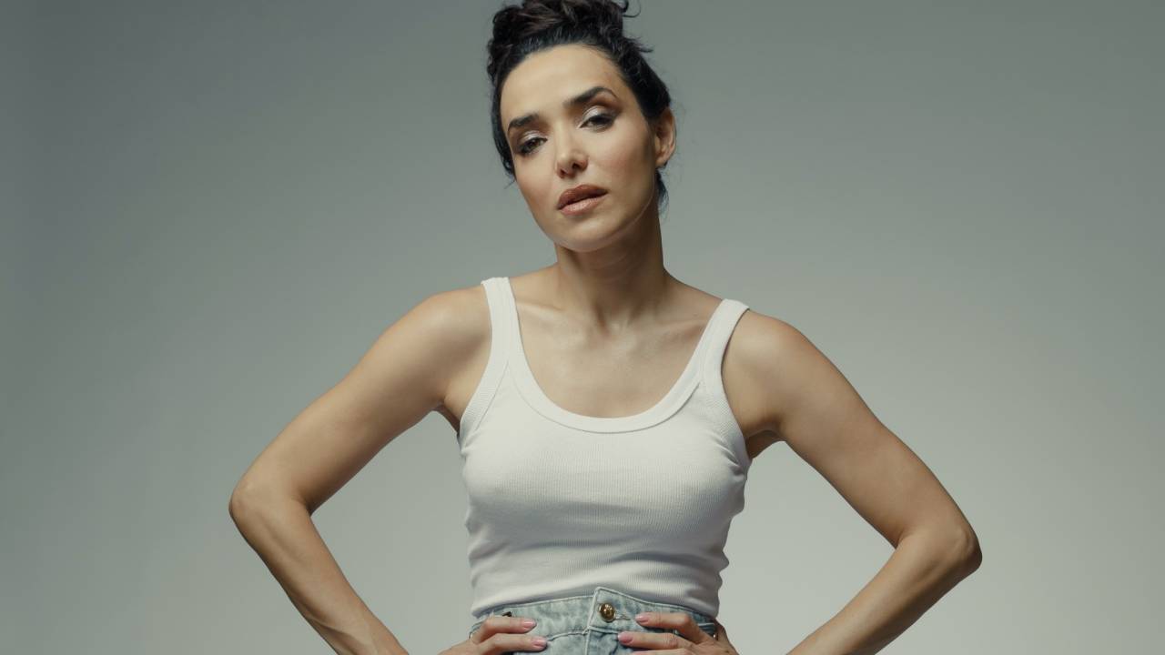 Mulher branca posa com as mãos na cintura e coque na cabeça. Veste regata branca e jeans clara.