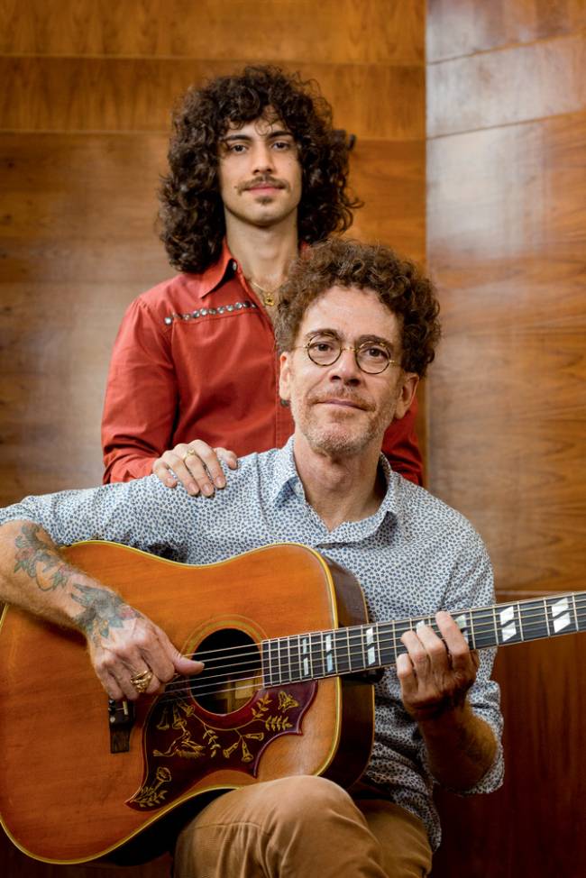 nando reis e sebastião Imagem mostra Nando sentado, segurando violão e, atrás do cantor, o filho em pé, com a mão no ombro do pai