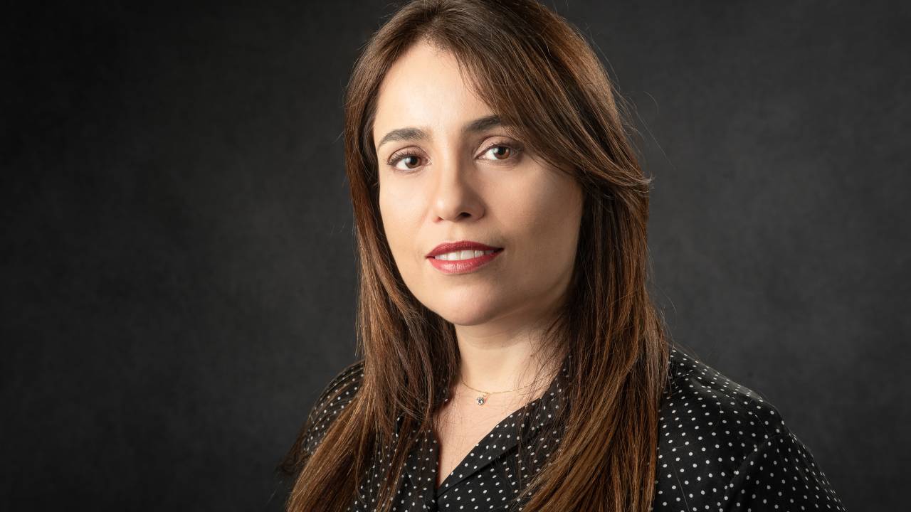 Mulher branca de cabelos lisos longos e castanhos encara a câmera. Veste camisa preta.