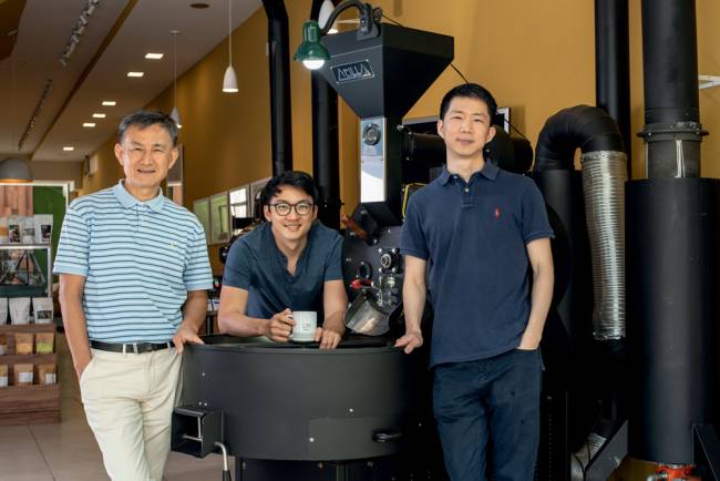 Boram, Garam e Stefano Um – Um Coffee Co. Imagem mostra pai e dois filhos em cafeteria, apoiados em equipamento grande de café. Eles estão em uma cafeteria, posicionados lado a lado e sorriem para a foto