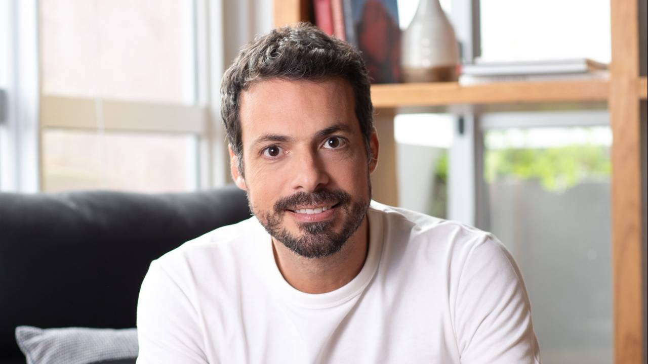 Imagem mostra João sentado em sofá de sala iluminada. Ele está com os braços cruzados em cima das pernas, usa camiseta branca e calça jeans e sorri