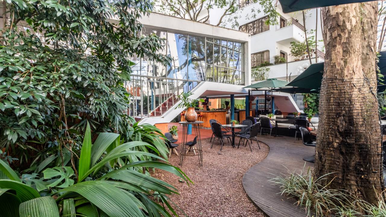 Jardim à esquerda esconde parte de grande casa branca envidraçada, com área aberta e mesas.