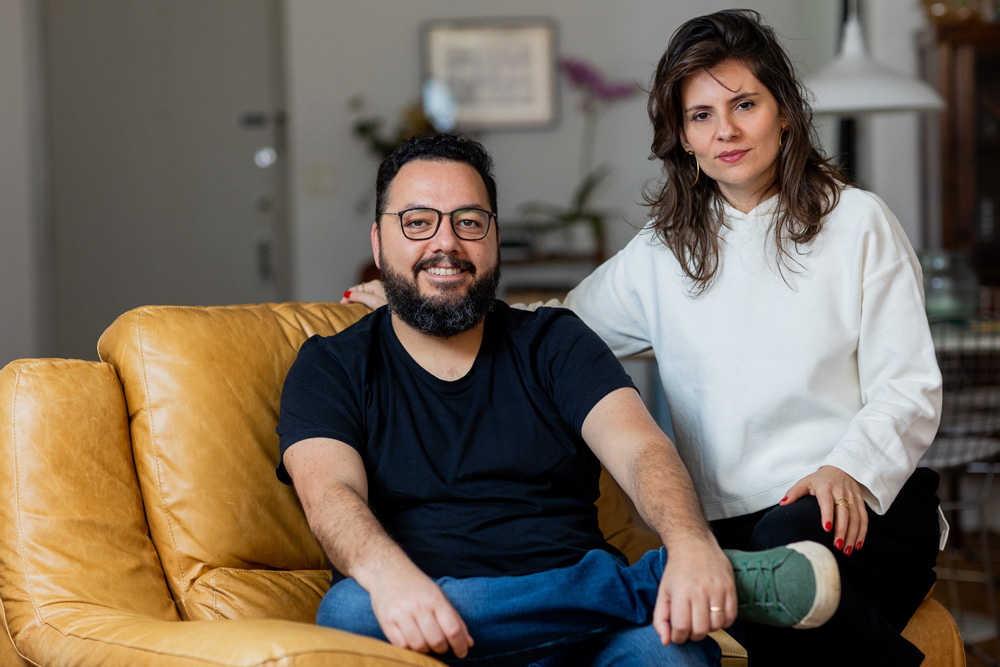 Homem com barba e óculos sorri ao lado da esposa, de cabelos lisos castanhos. Ele veste camiseta preta e jeans, ela um suéter branco. Eles estão em uma sala de estar com poltrona de couro claro, quase amarelado.