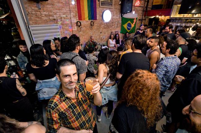 Daniel olha para a foto segurando um drinque em ambiente lotado com roda de samba ao fundo