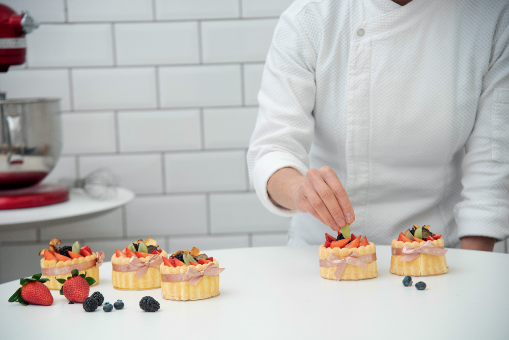 COMER & BEBER 2023 – Amay Patisserie Sobre uma mesa branca tem tortinhas com frutas em cima. Ao fundo, uma pessoa de avental branco coloca uma uva sobre uma tortinha.
