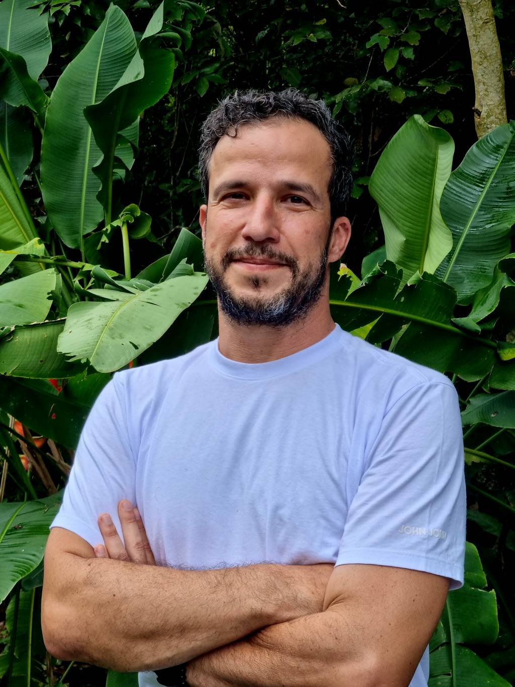 André Palhano (Zeca Prado-Divulgação) Homem branco de barba dá meio sorriso. Veste camiseta branca e posa em frente a área verde.