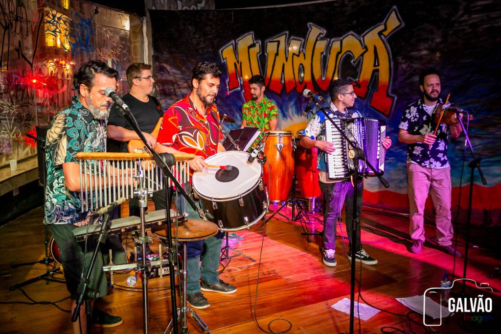 Banda Munducá (Ricardo Galvão-Divulgação) (1) Foto iluminada em tom vermelho exibe integrantes de banda de forró manipulando seus instrumentos musicais.