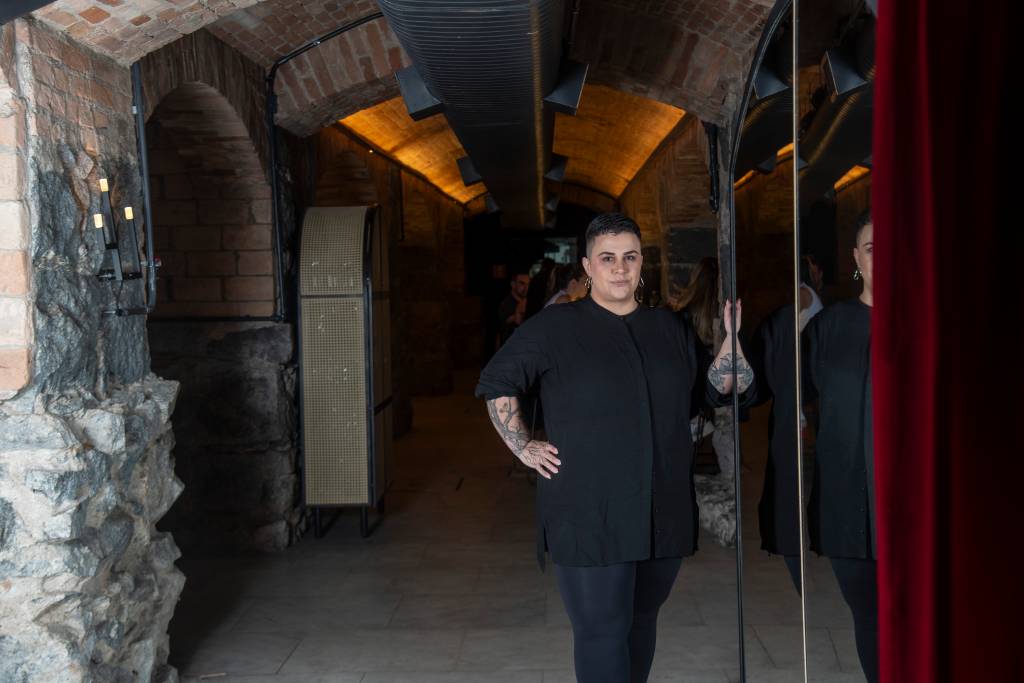 Bar dos Arcos Mulher toda de preto de cabelo curto com a mão direita na cintura.