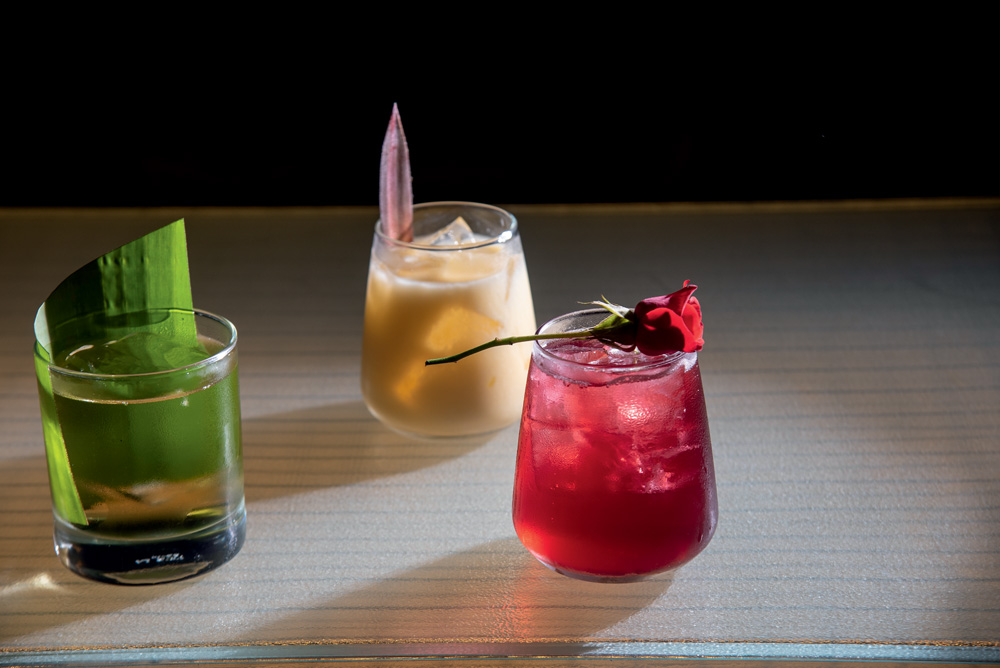 COMER & BEBER 2023 – Bar dos Arcos Sobre uma mesa branca, três drinques de cores diferentes. O da esquerda é translúcido, com uma folha verde dentro do copo. O do meio é bege. Mais à direita, um drinque rosa com uma flor deitada na borda do copo.