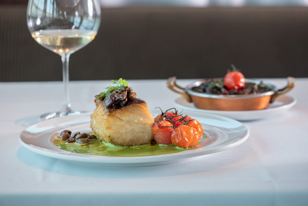 COMER & BEBER 2023 – Bistrot Parigi Prato branco com pedaço de bacalhau, legumes e molho de manjericão embaixo.