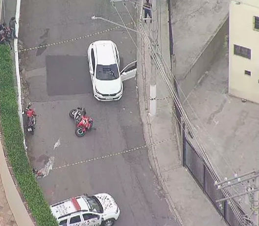 a imagem mostra um carro branco com marcas de tiro no para-brisa e, ao lado, uma moto caída no chão