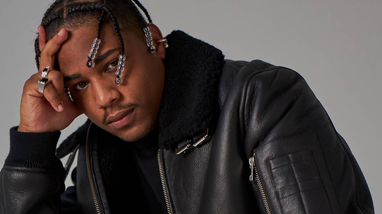 Homem negro posa com mão na cabeça. Ele tem dreads com detalhes coloridos e veste jaqueta preta de couro.