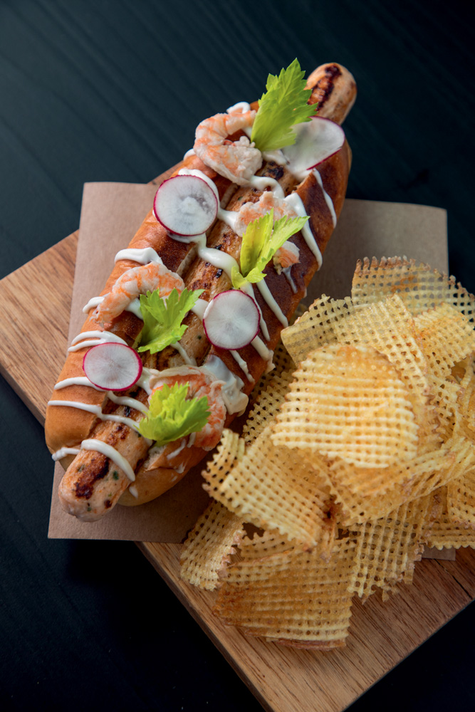 COMER & BEBER 2023 – Astor Uma tábua de madeira com batatas chips de um lado e do outro um hot dog com pedaços de camarão por cima e algumas folhas verdes.