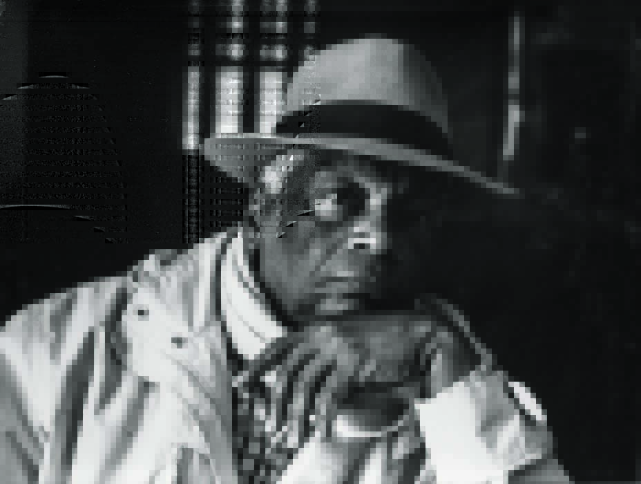 Foto em preto e branco de um homem negro, usando chapéu, posando com semblante neutro