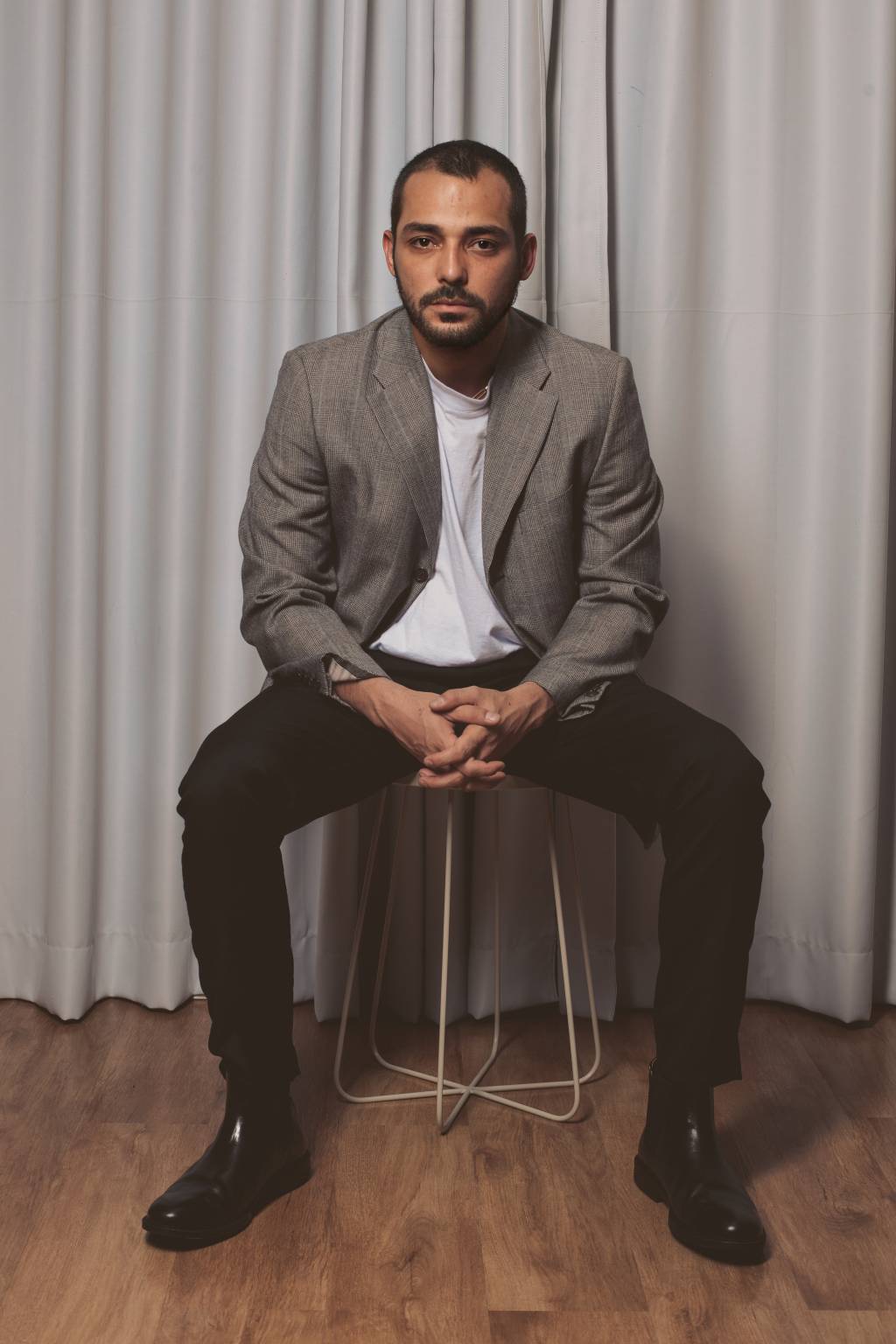 Edu Sterblich (Thalles Leamari-Divulgação) Homem branco de barba rala posa com olhar sério em banquinho, com as os dedos unidos e pernas abertas. Veste blazer cinza, camiseta branca e calça preta.