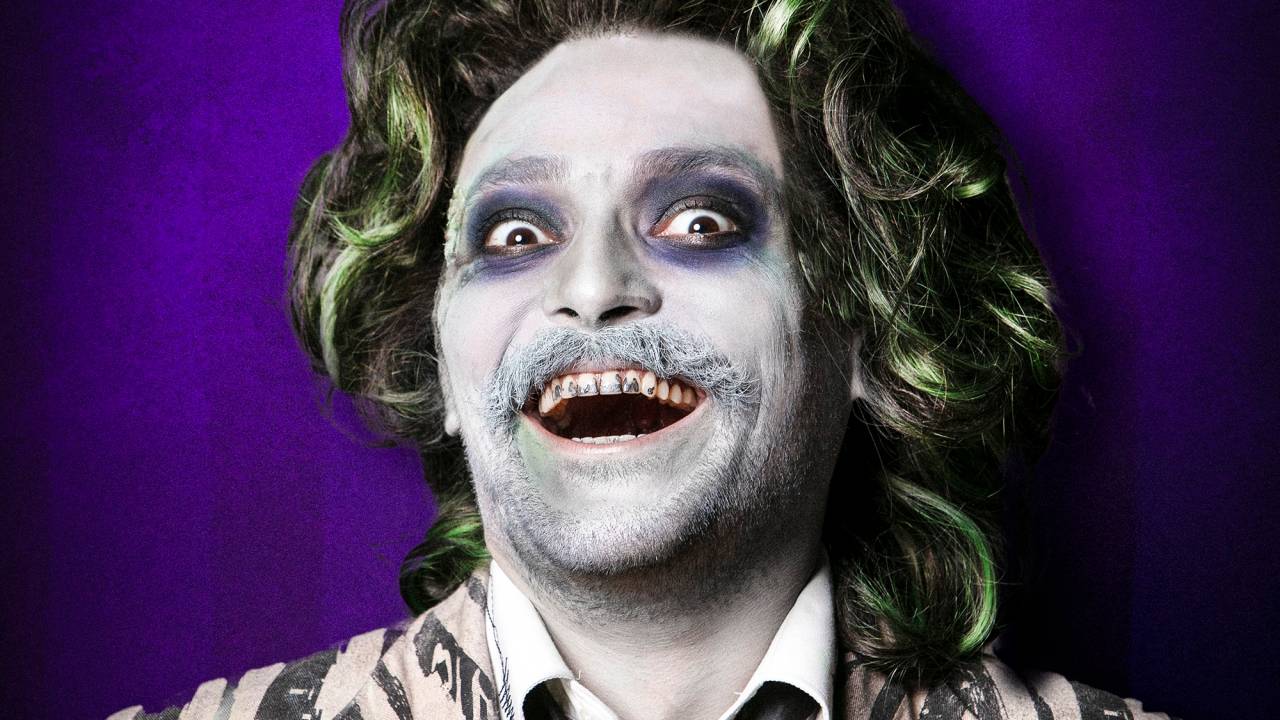 Em fundo roxo, homem todo maquiado de branco posa em meio sorriso assustador e irônico. Os cabelos são volumosos e levemente esverdeados. O blazer tem listras pretas e brancas e os olhos fundos têm círculos pretos.