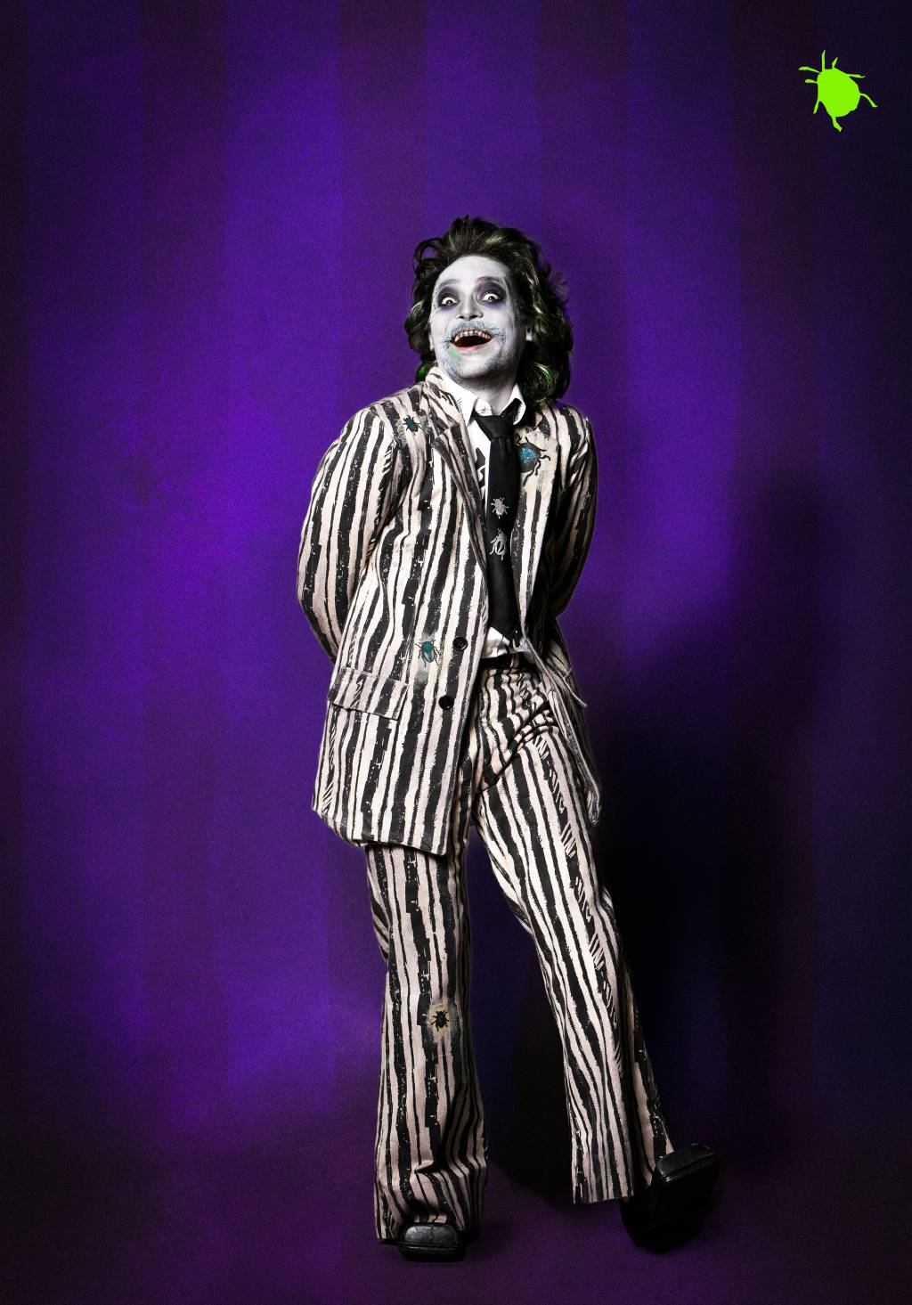 Eduardo Sterblitch como Beetlejuice (Leo Aversa-Divulgação) (3) Em fundo roxo, homem todo maquiado de branco posa em meio sorriso assustador e irônico. Os cabelos são volumosos e levemente esverdeados. O blazer tem listras pretas e brancas e os olhos fundos têm círculos pretos. As mãos estão escondidas atrás das costas.