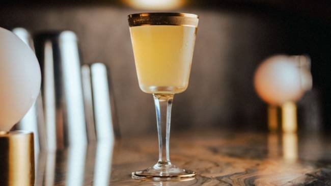 A imagem mostra uma taça de vidro com detalhe de cor cobre nas bordas e um líquido amarelo claro. A taça está em cima de uma mesa de madeira.