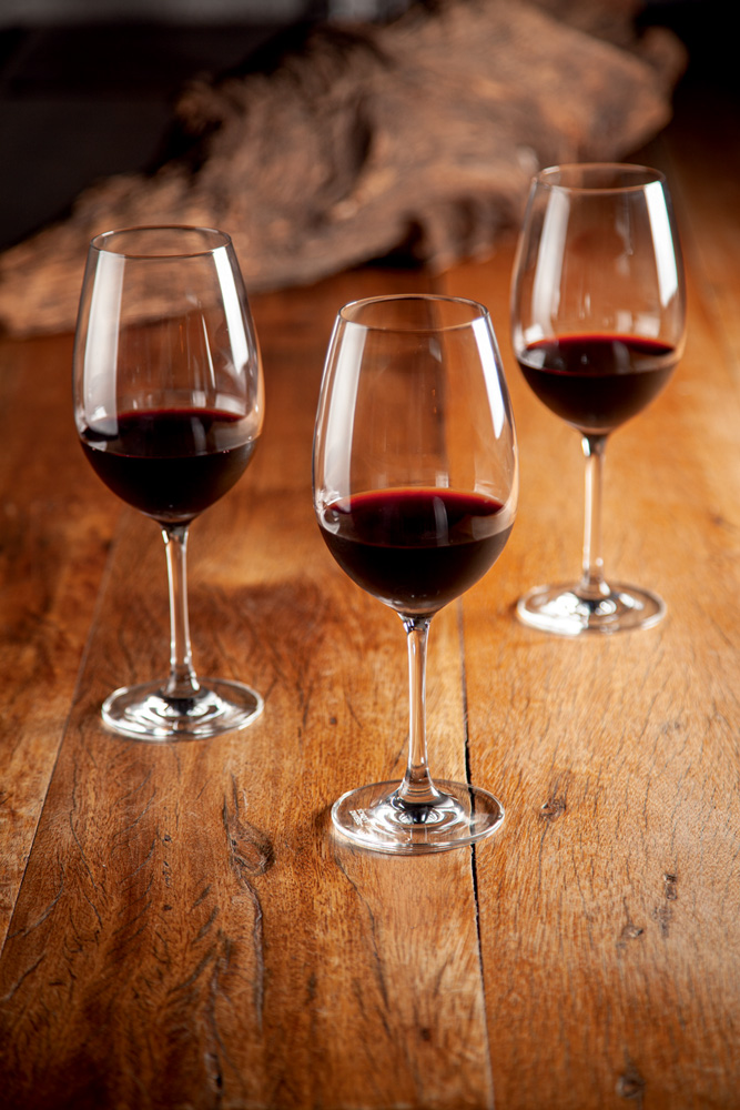 COMER & BEBER 2023 – Elevado Bar Três taças com vinho tinto sobre uma mesa de madeira.