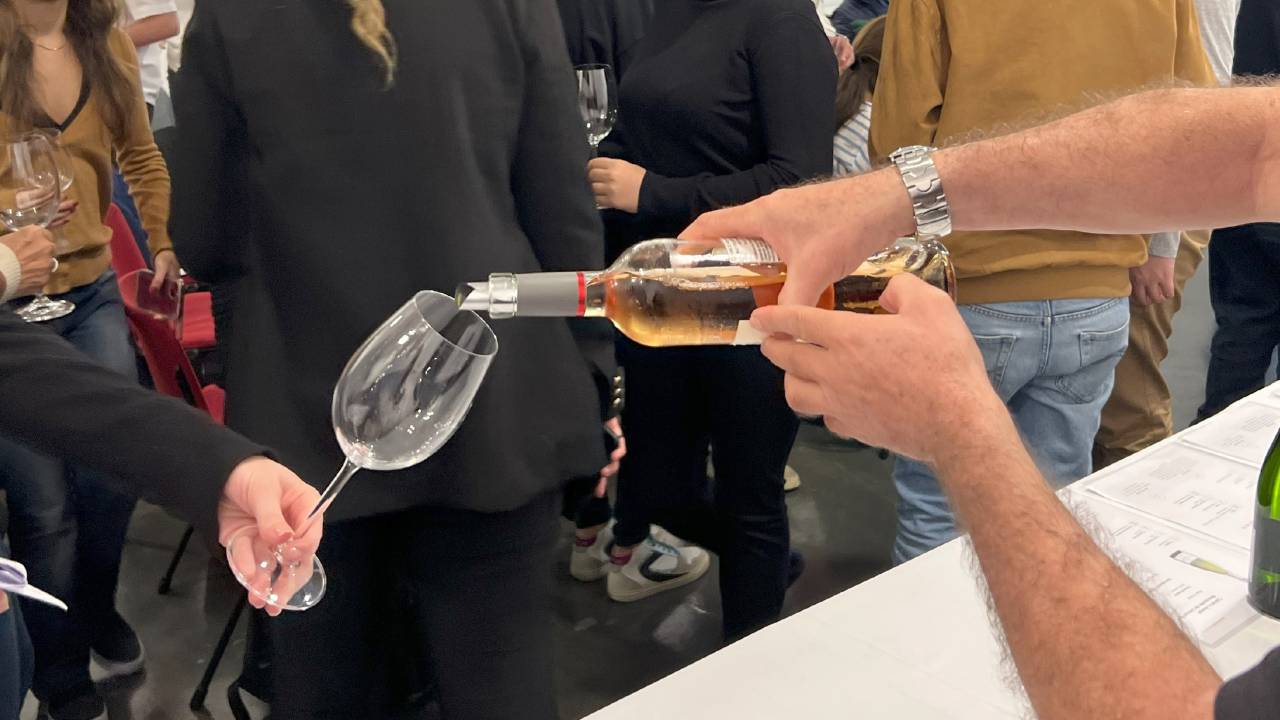 A imagem mostra duas pessoas segurando taças de vinho vazias, enquanto outra pessoa coloca o vinho.