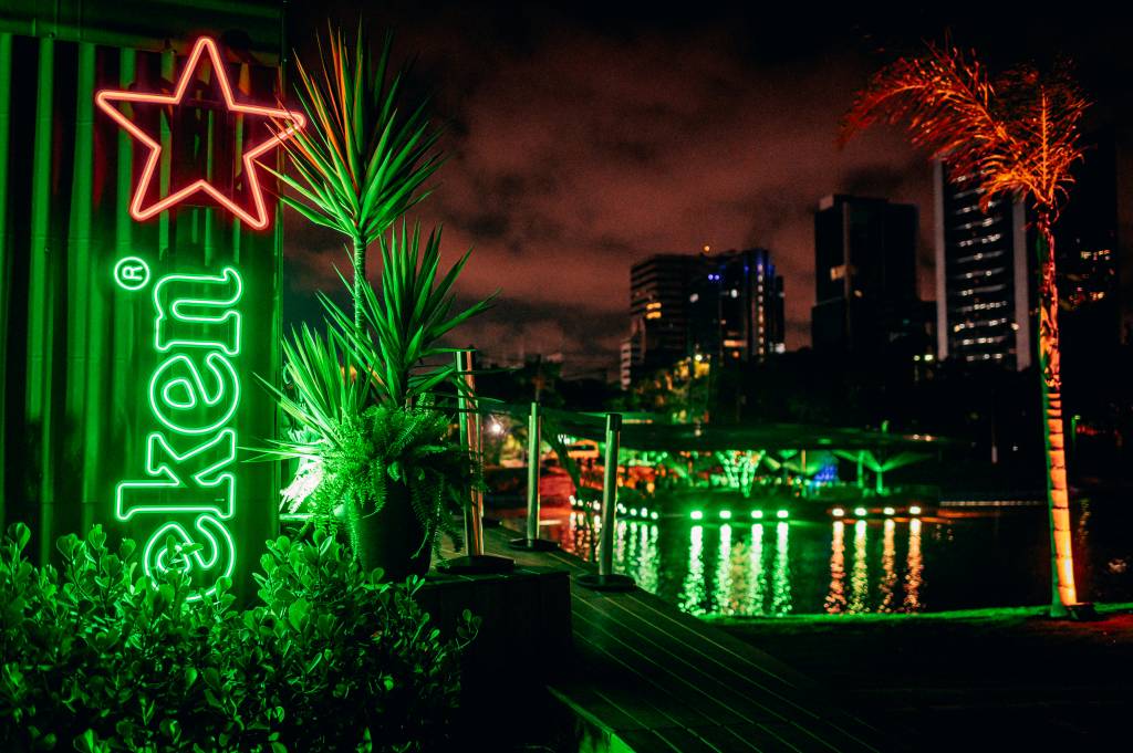 Heineken Floating Bar – Helena Yoshioka (1) Imagem destaca bar em luz verde no meio de rio e prédios iluminados à noite de fundo.