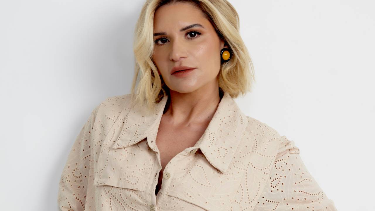 Mulher loira de cabelos curtos posa com a mão na cintura em fundo branco. Veste saia estampada e camisa bege.