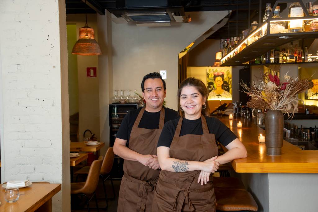 Metzi Homem, à esquerda, e mulher, à direita, de camiseta preta e avental marrom em salão de restaurante.