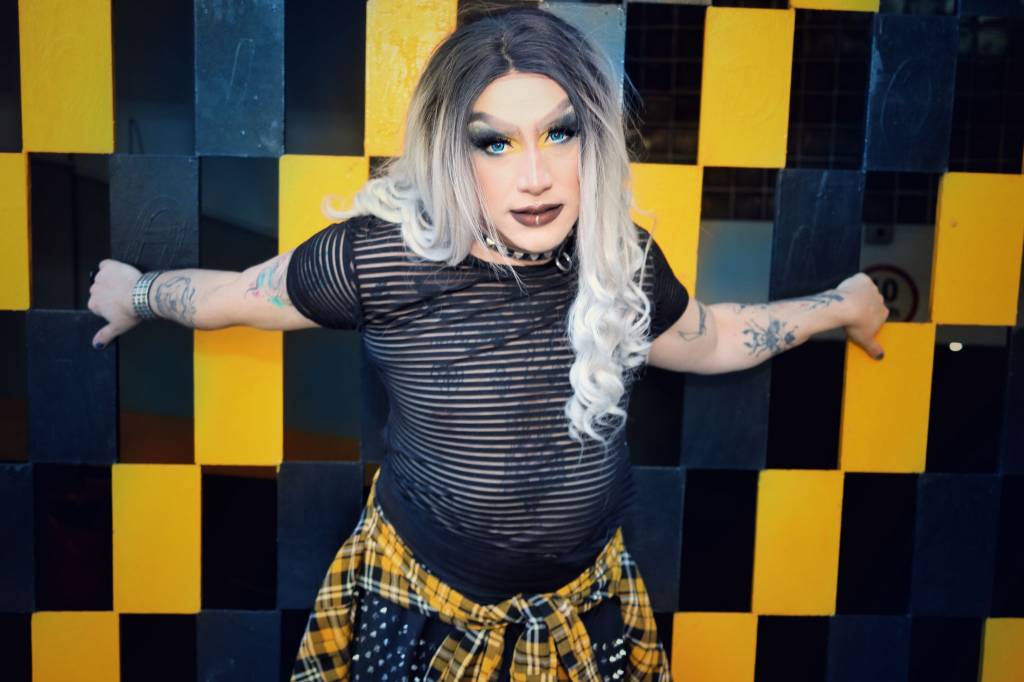 Drag queen posa com os braços na parede quadriculada, usa peruca de cabelo loiro longo e veste camiseta preta transparente com jaqueta quadriculada na cintura.