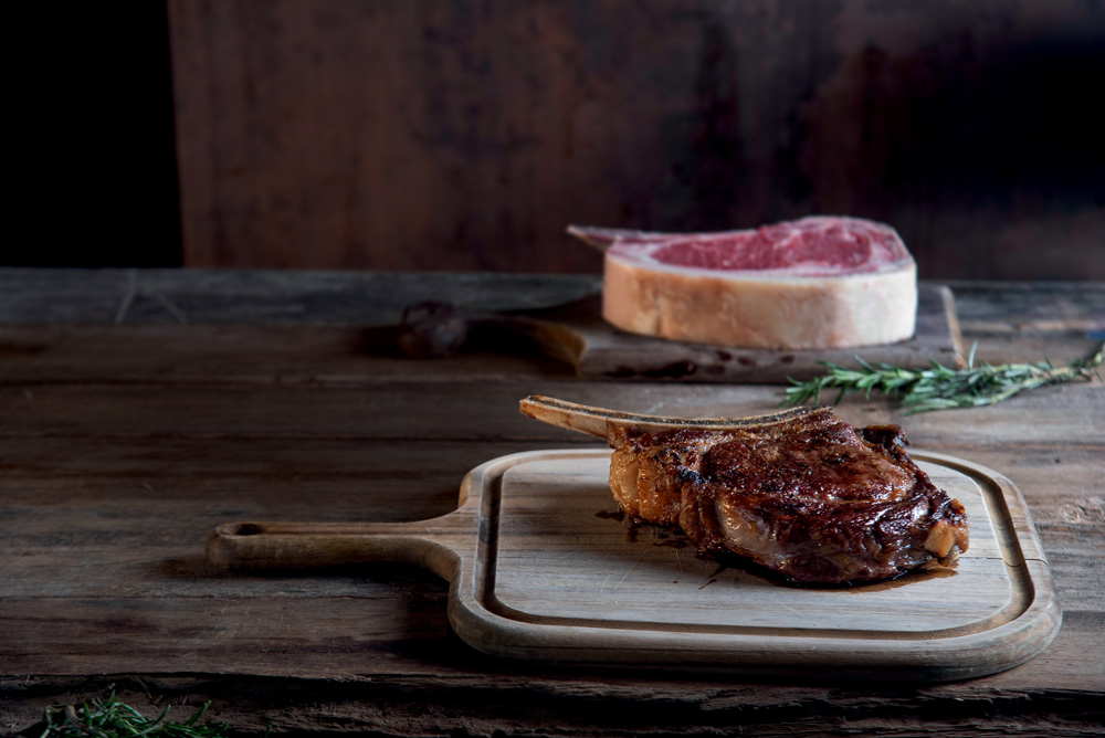 COMER & BEBER 2023 – Osso Imagem mostra dois pedaços de carne com osso, um está passado e dourado, o outro ao fundo está cru.