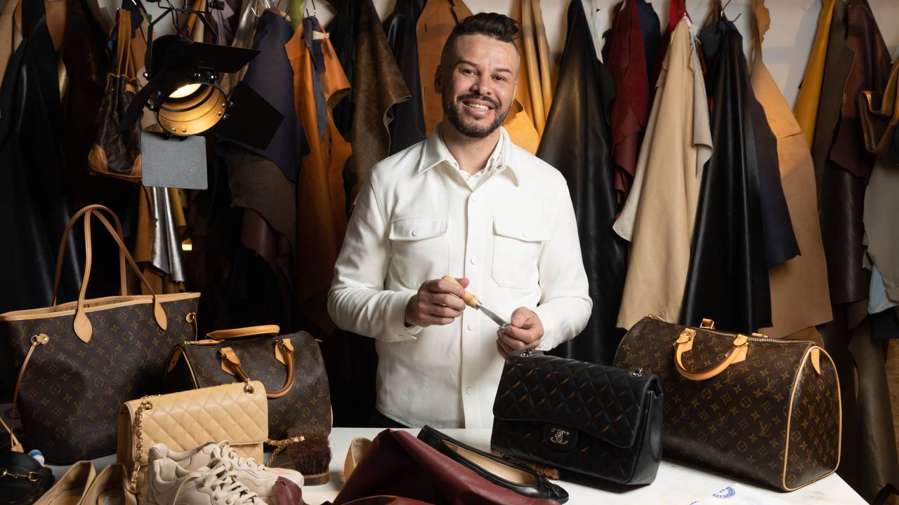 Homem com barba posa segurando instrumento pontiagudo prata em frente a mesa de sapateiro com bolsas de grife em exibição.