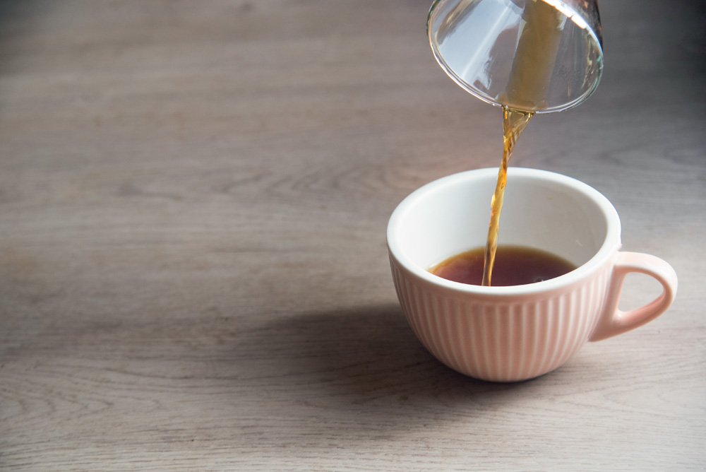 COMER & BEBER 2023 – Um Coffee Co. Café coado sendo despejado em uma xícara branca sobre uma mesa de madeira.